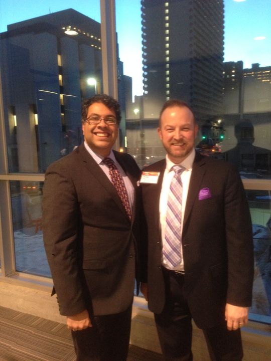 Michael Bach with Naheed Nenshi, former Mayor of Calgary, at an evening event in downtown Calgary with the city skyline visible through the window behind them.